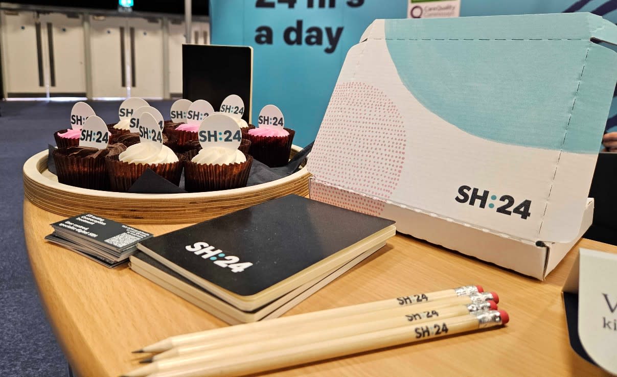 A table set out with SH:24 branded items, including pencils and notebooks. In the background, you can see cupcakes decorated with the SH:24 logo.