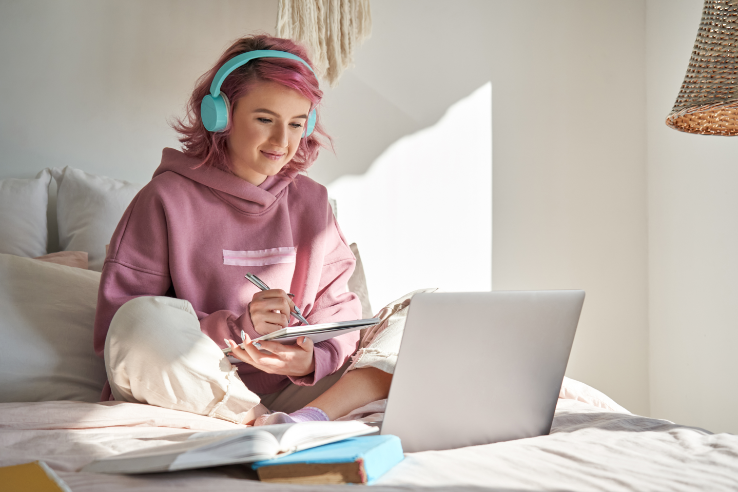 Junge Frau sitzt mit Laptop und Schreibeutensilien auf ihrem Bett 