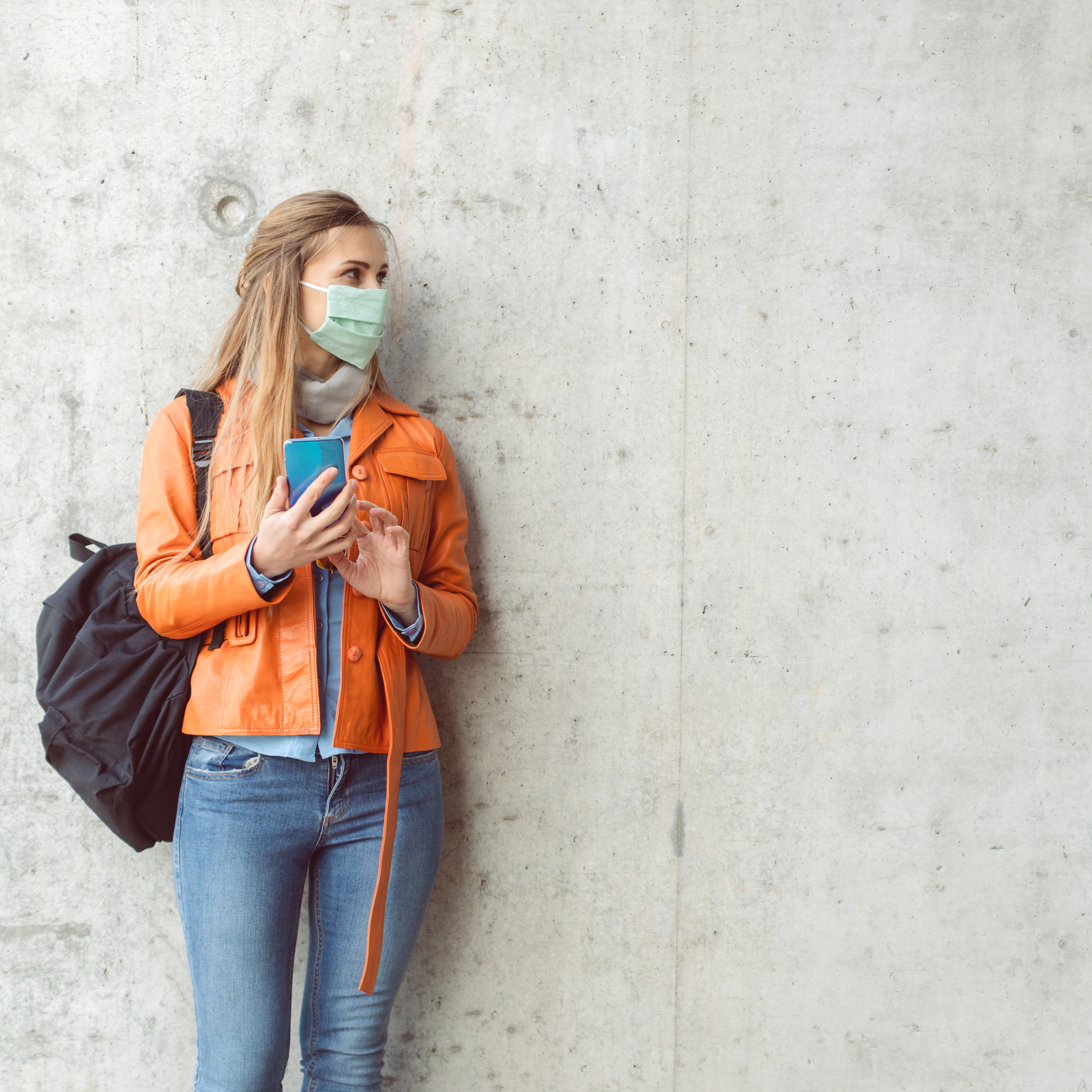 Eine Frau die ein Handy in der Hand hat, eine Maske trägt und in die Ferne schaut