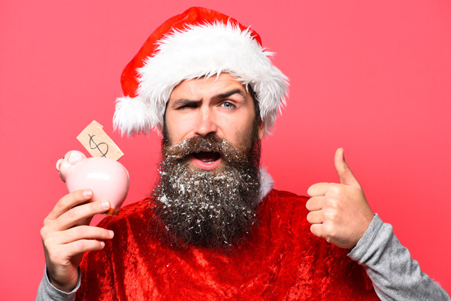 Mann mit Weihnachtsmütze schaut in die Kamera und hält den Daumen hoch und in der anderen Hans ein Sparschwein