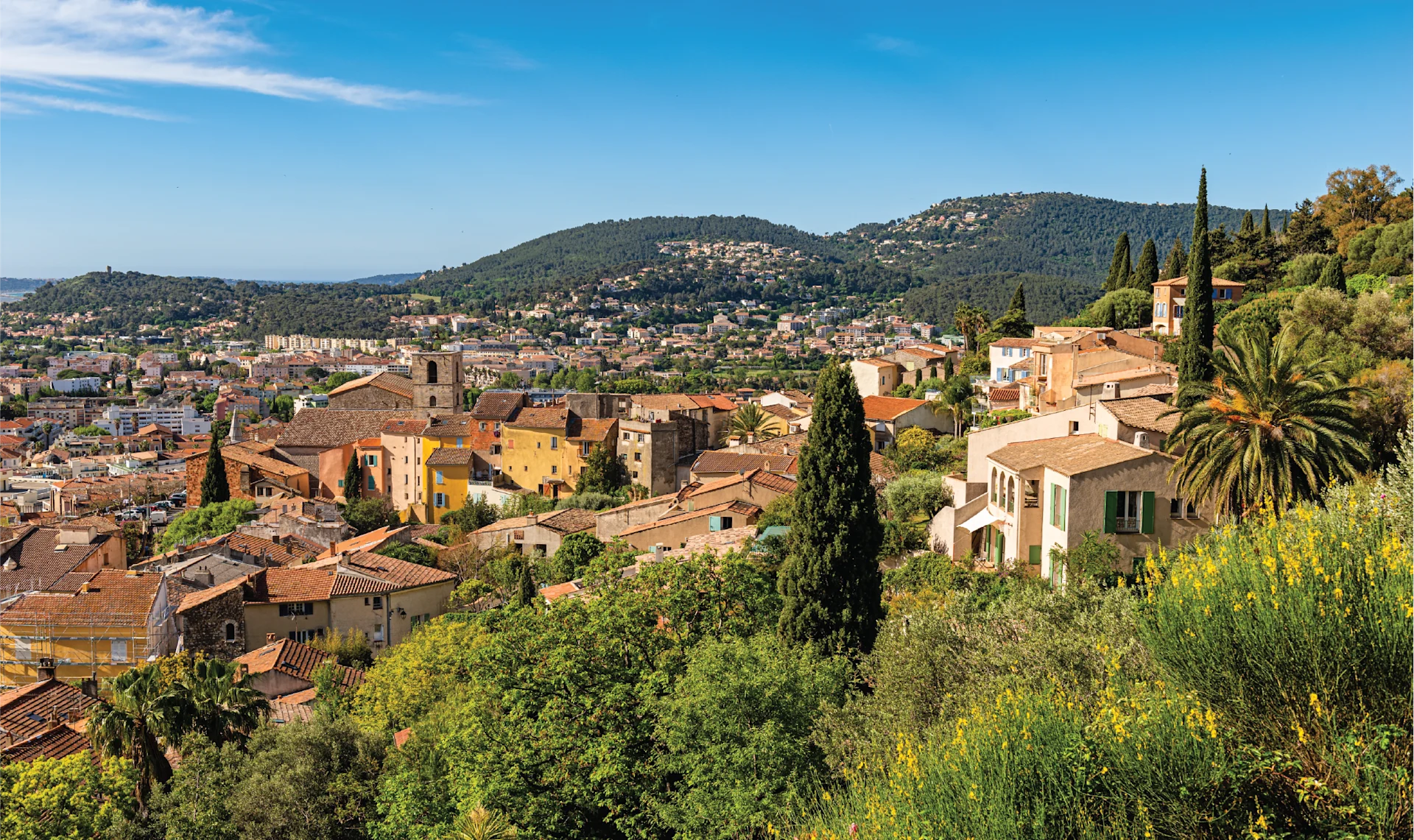 Panoramic view over a coastal Mediterranean town of Hyères with terracotta rooftops, rolling green hills, cypress trees, and a clear blue sky.