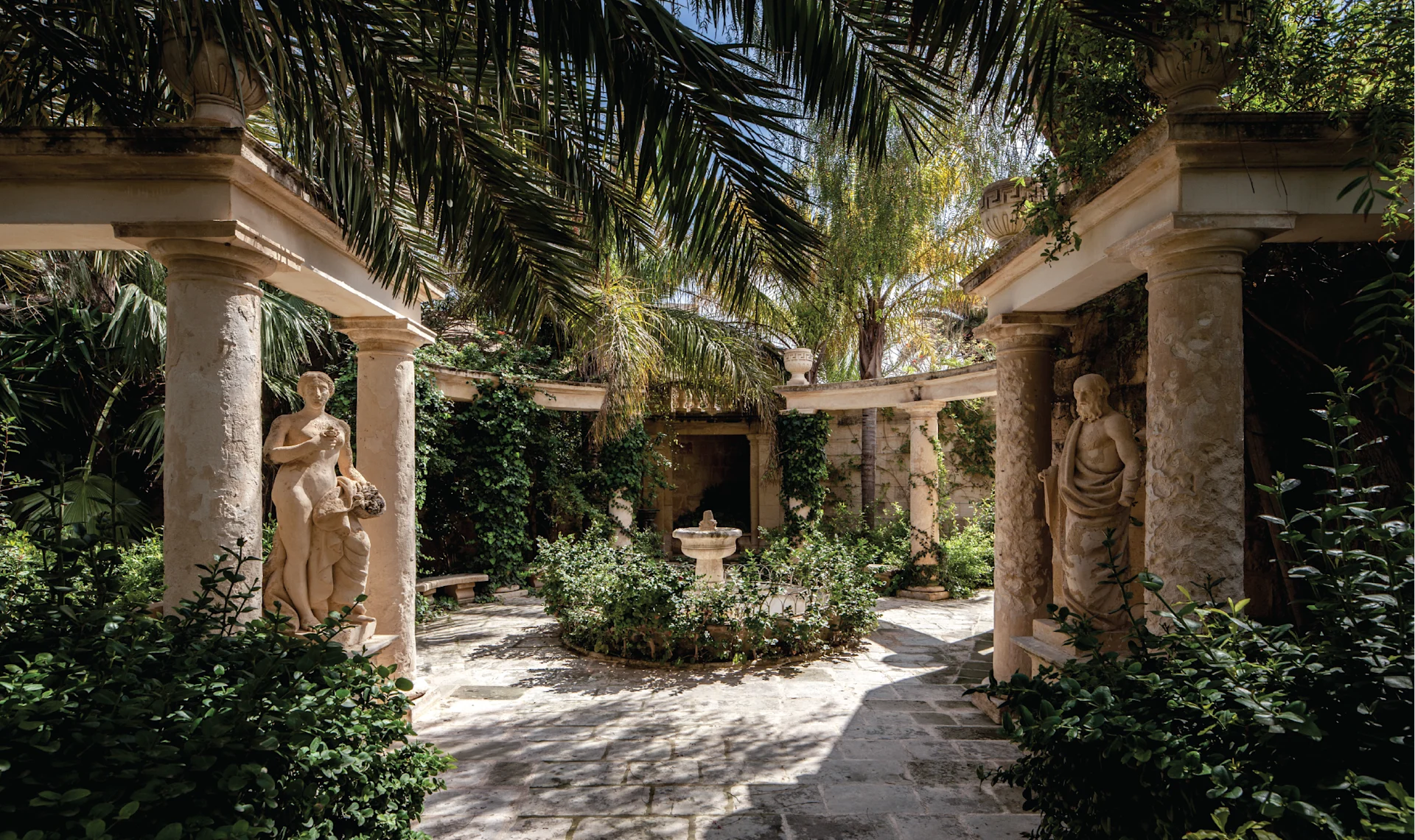 Secluded garden courtyard at Casa Bonavita with classical statues, stone colonnades, lush greenery, and a central fountain creating a peaceful oasis.