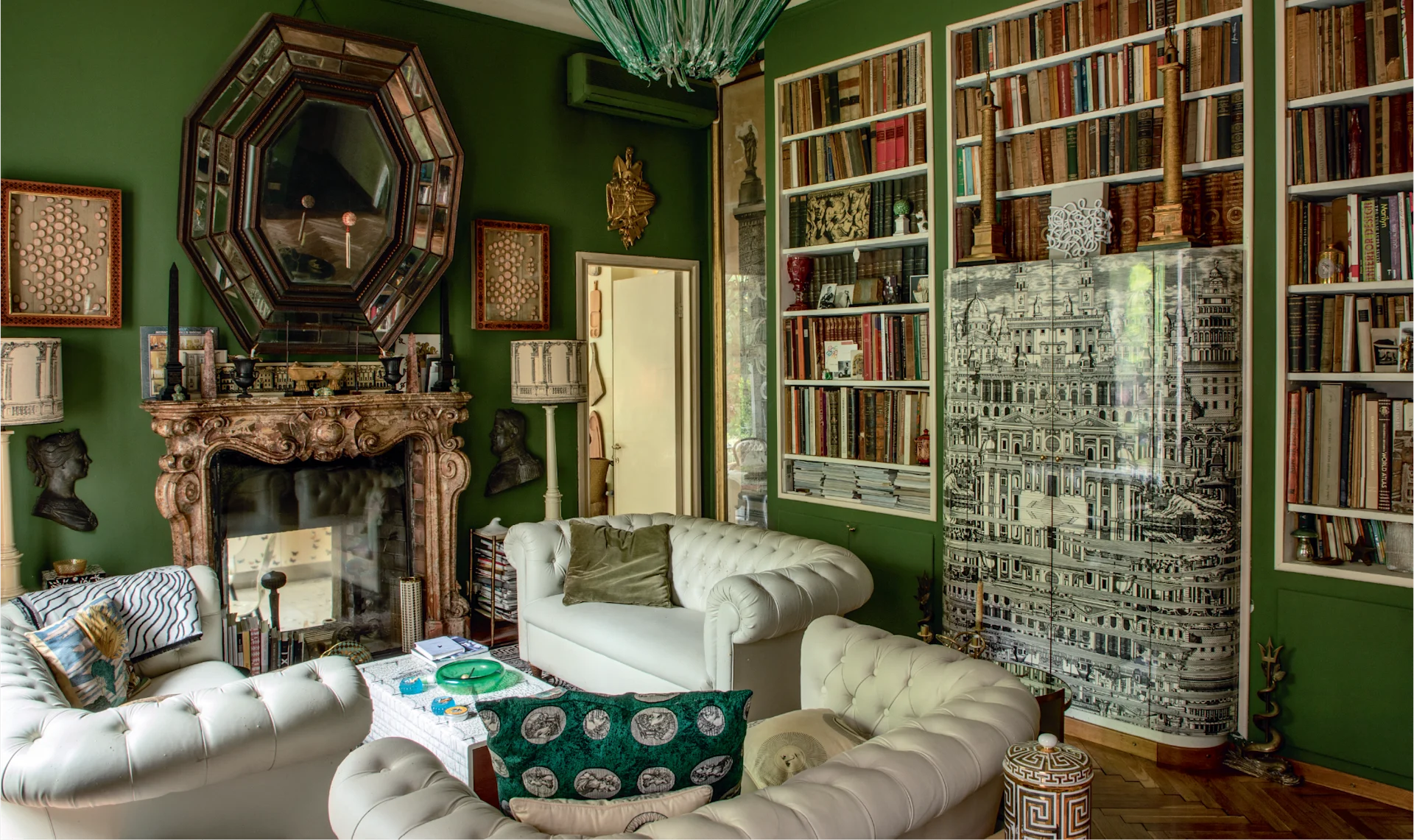 Luxurious library painted deep green with floor-to-ceiling bookshelves, a marble fireplace, sculptural mirrors, and plush white sofas arranged around a low table.
