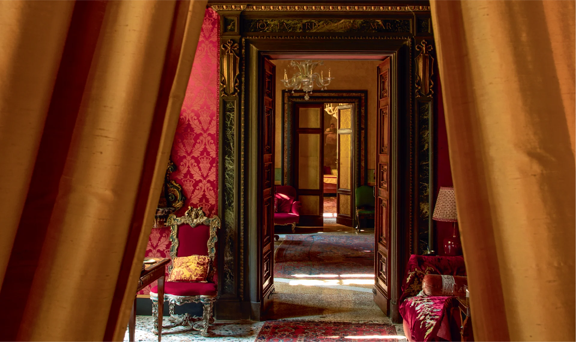 Dramatic interior view framed by golden curtains, revealing a richly decorated room with deep red walls, ornate carpets, antique furniture, and layered doorways creating a sense of depth.