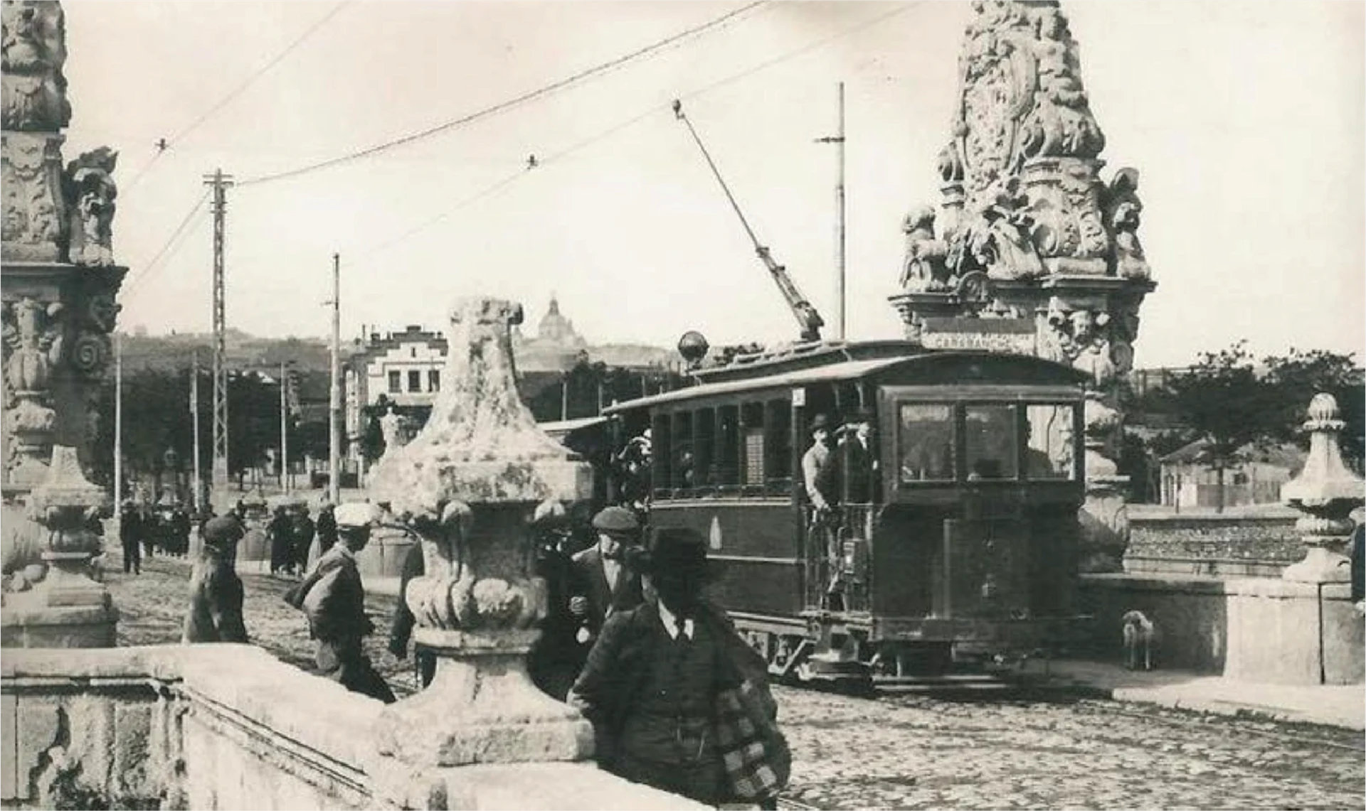Historic black-and-white photograph of a tram crossing Puente de Toledo in Madrid in 1912, with pedestrians standing along the bridge’s sculpted balustrade.