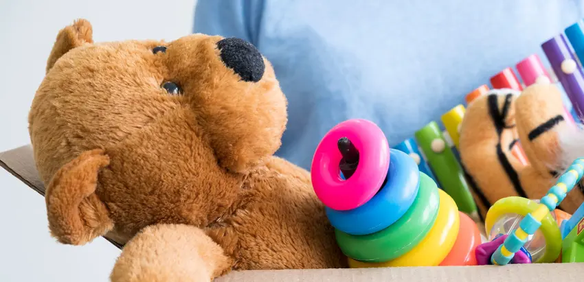 Caixa de cartão com um urso de peluche castanho, um xilofone colorido, um jogo de empilhar e partes de outros brinquedos de bebé.