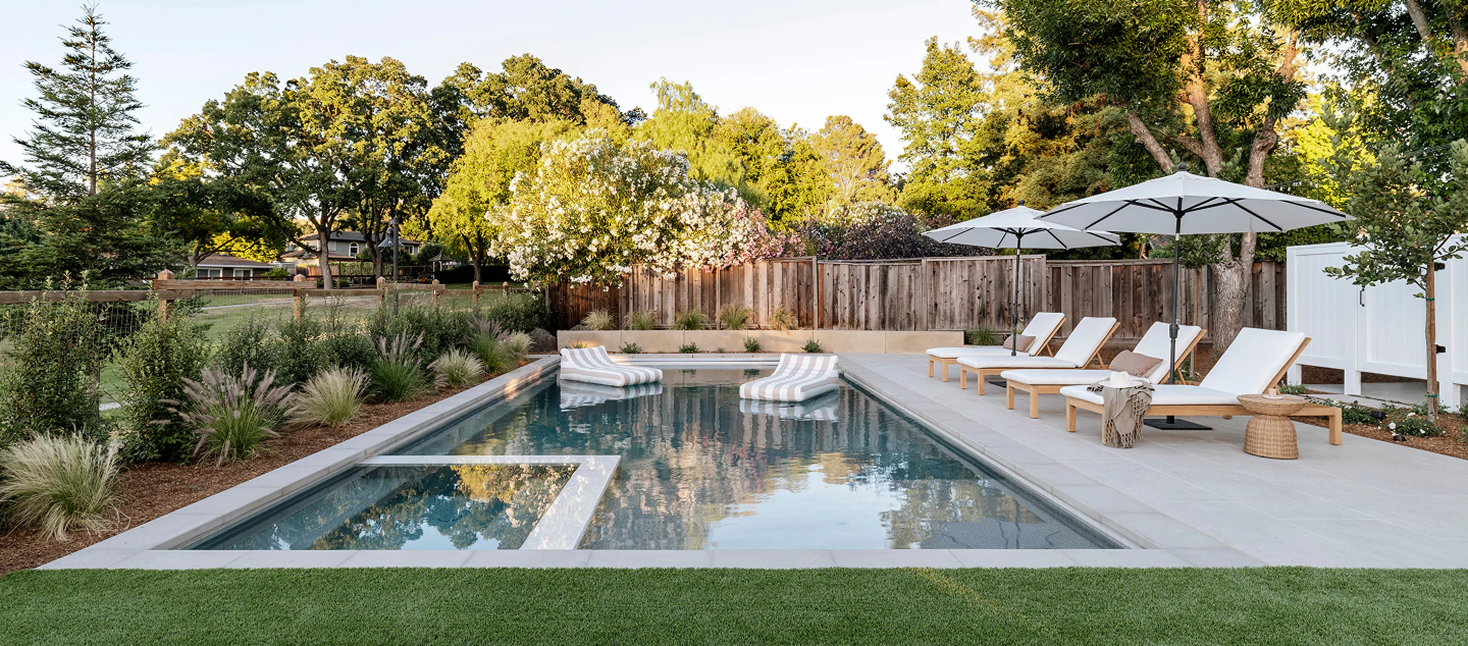 Contemporary backyard pool with in-water loungers, white poolside chairs, and large umbrellas framed by lush landscaping.