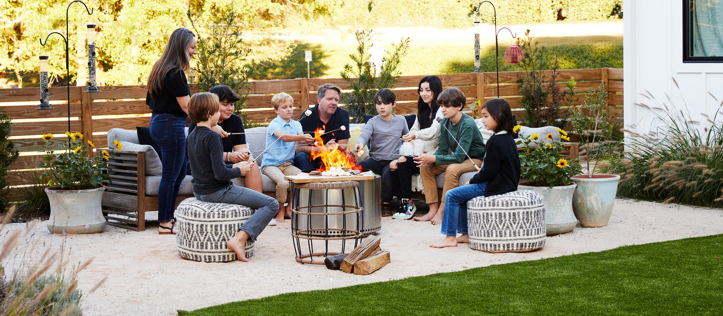 Group of people gathered around a firepit in a modern outdoor living space, surrounded by comfortable seating and landscaping.