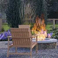Wooden outdoor chair facing a round stone firepit with flames, surrounded by plants and a wooden fence.