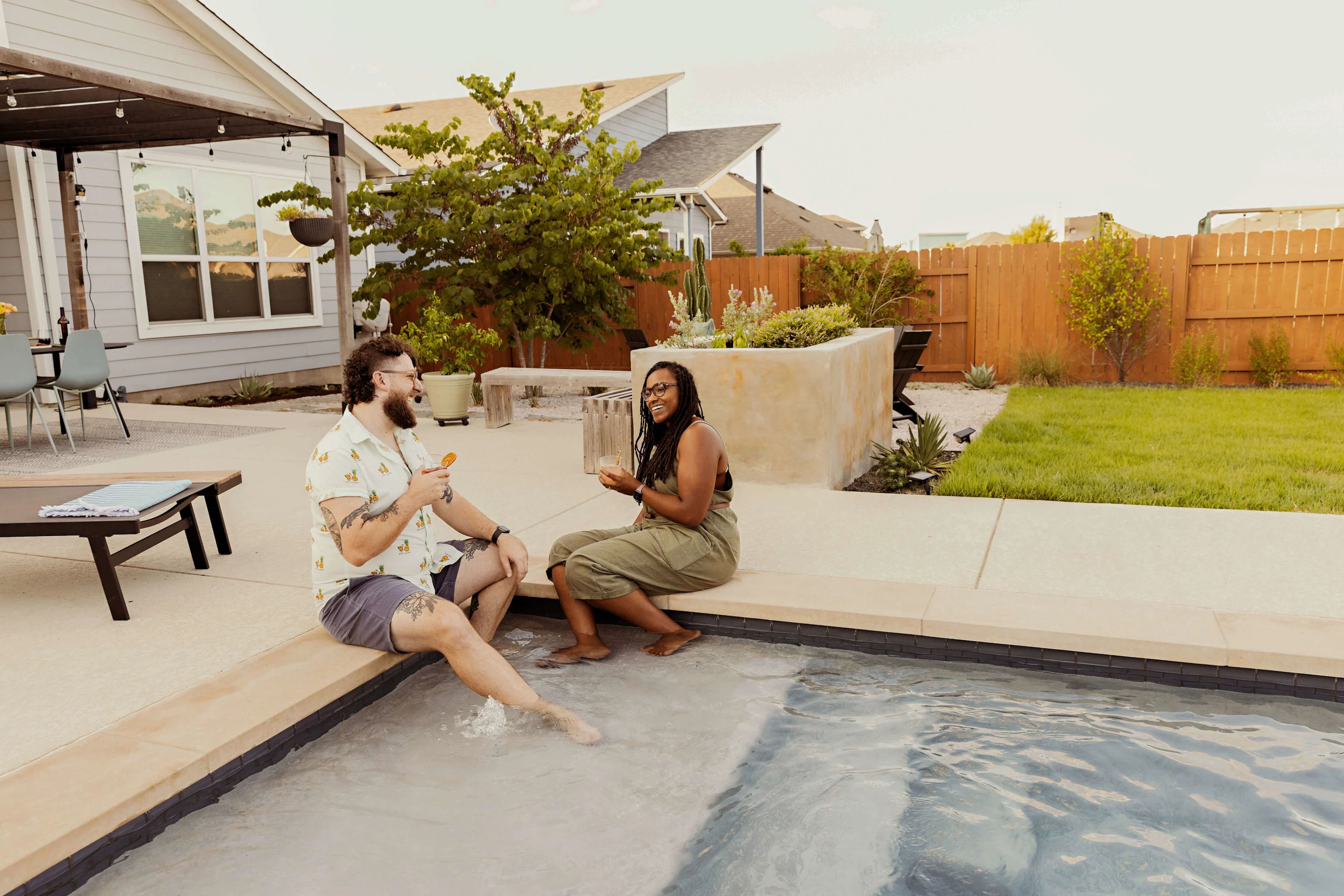couple sitting at the edge of swimming pool in back yard.