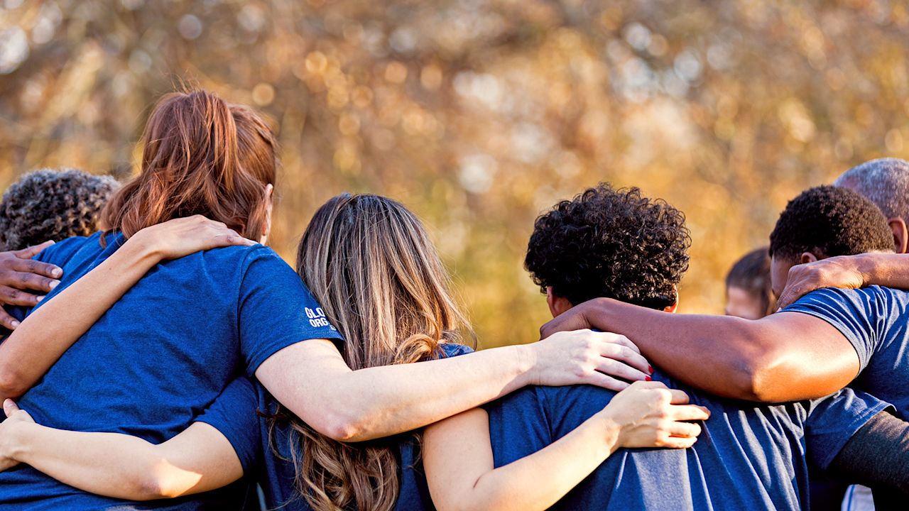 Eine Gruppe von Menschen bilden einen Kreis, alle legen ihre Hände über die Schultern der Personen neben ihnen.
