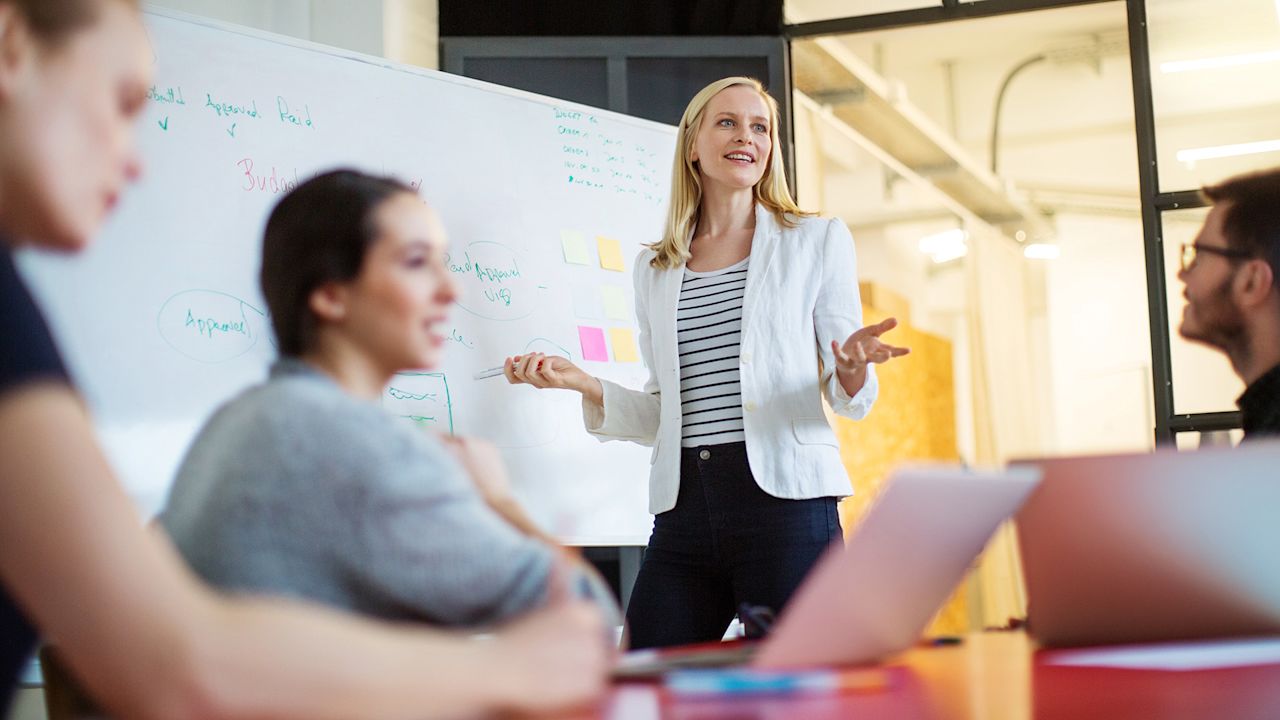 Eine Frau steht vor einer Gruppe von Kollegen an einem Whiteboard und moderiert ein Meeting.