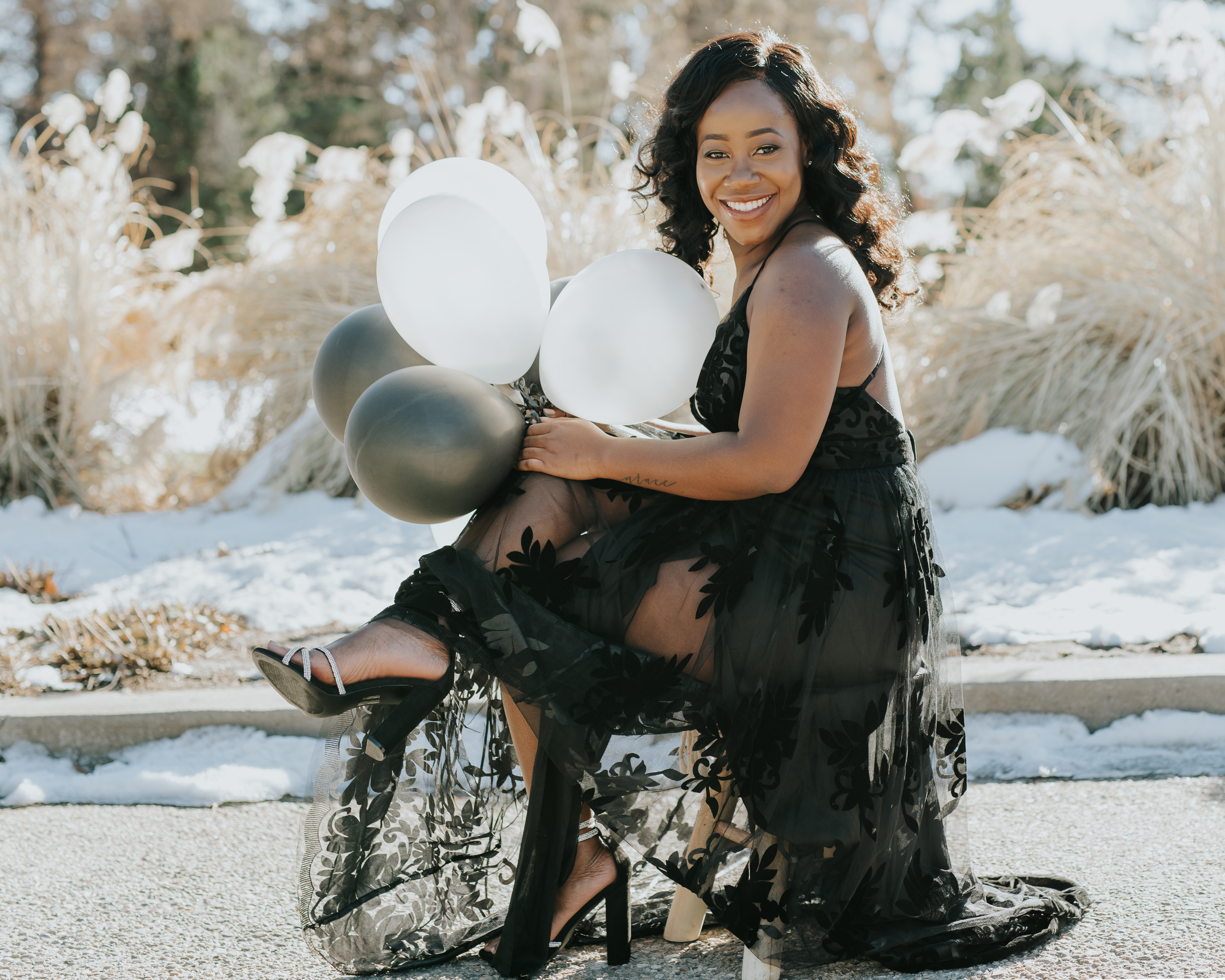 Woman in a black floral tulle gown sitting and laughing while holding a bundle of white and black balloons, another look captured during the same Shoott session.