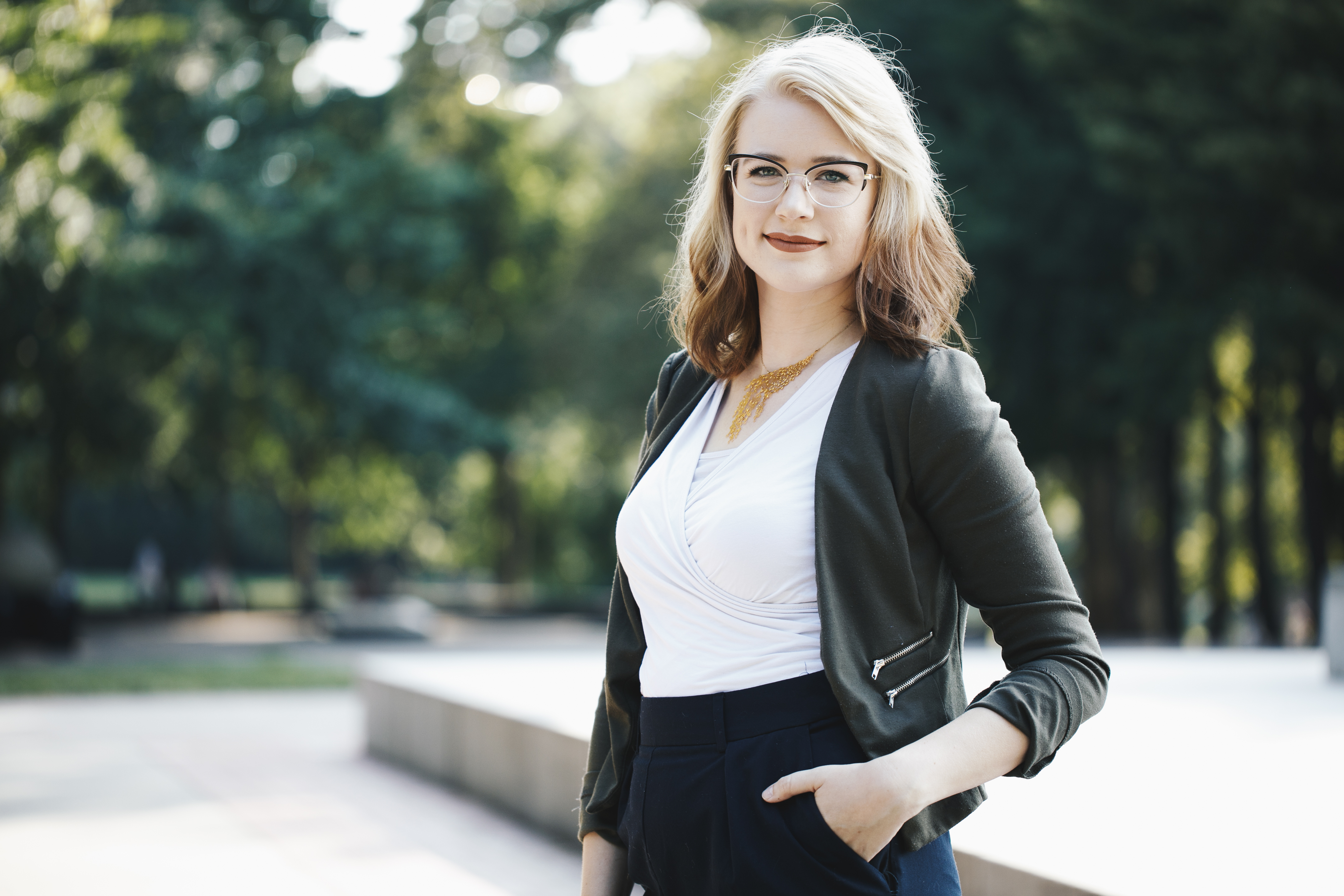Young woman with blonde hair and glasses smiling outdoors in a dark jacket and white top, hand in pocket, blurred green park background, San Antonio professional headshot by Shoott in natural light
