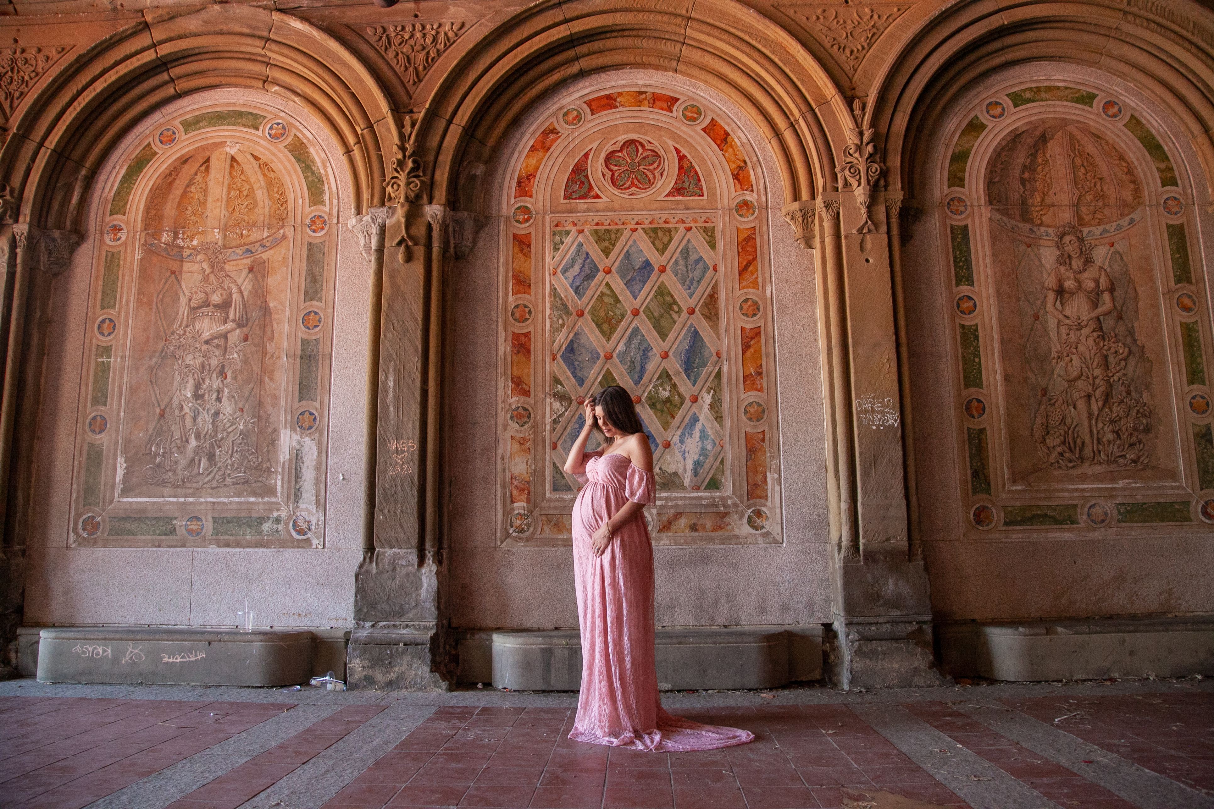 Pregnant woman in a pink off-shoulder lace gown standing under the ornate tiled arches of Bethesda Terrace in Central Park, captured during a Shoott maternity photoshoot