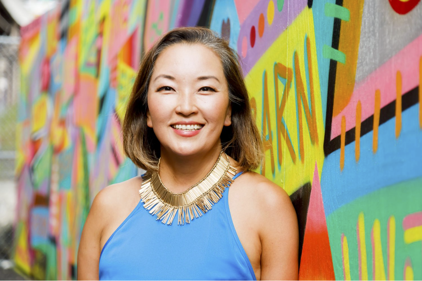 Woman in blue top and gold necklace smiling against a colorful graffiti wall during free professional portrait photo session with Shoott in San Antonio    