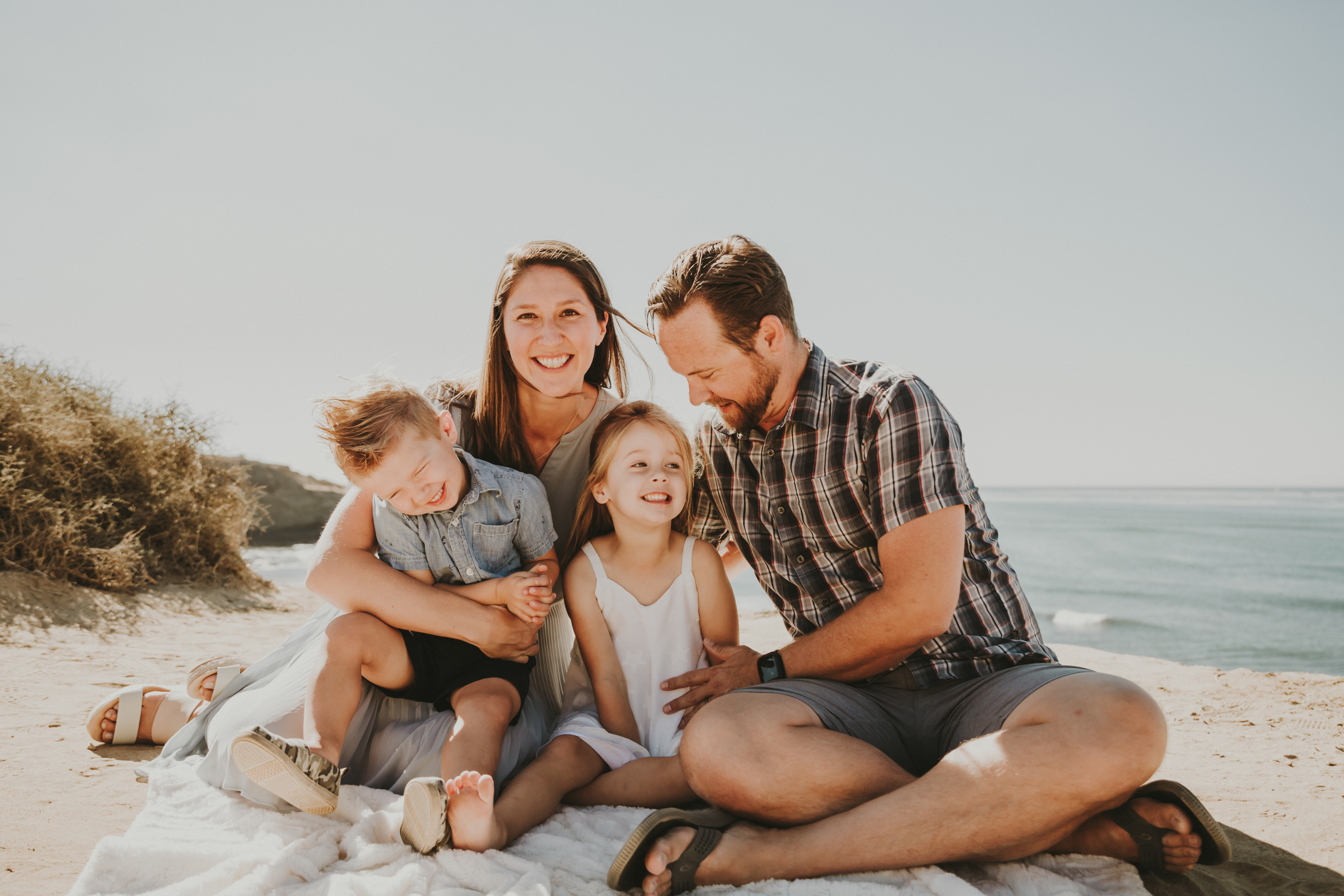 Family of four sitting together on a blanket on the beach, laughing and smiling at sunset by the ocean, captured during a Shoott family photoshoot.