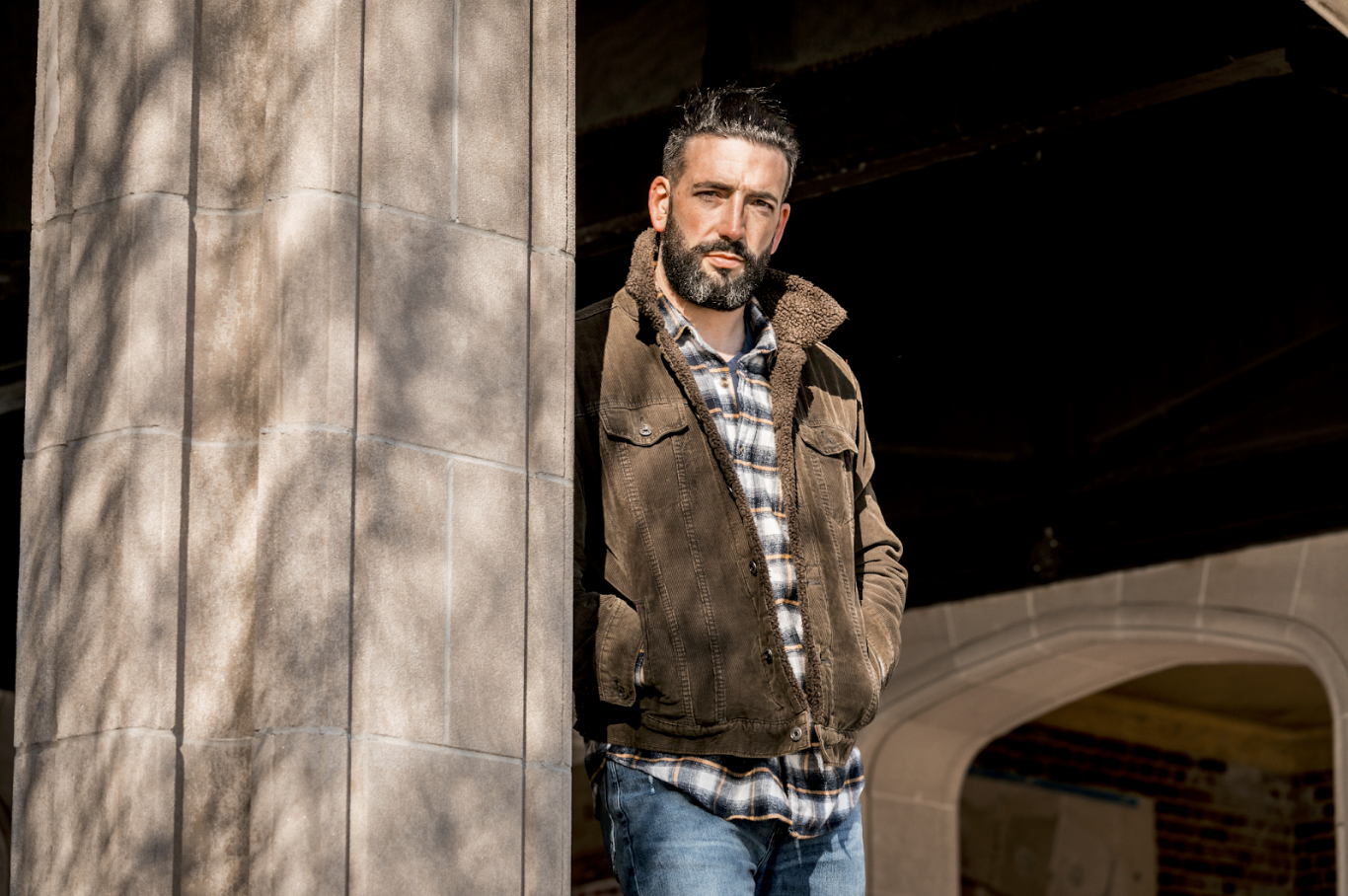 Man in suede jacket and plaid shirt leaning against a stone column in golden light during free professional lifestyle photo session with Shoott in Houston  