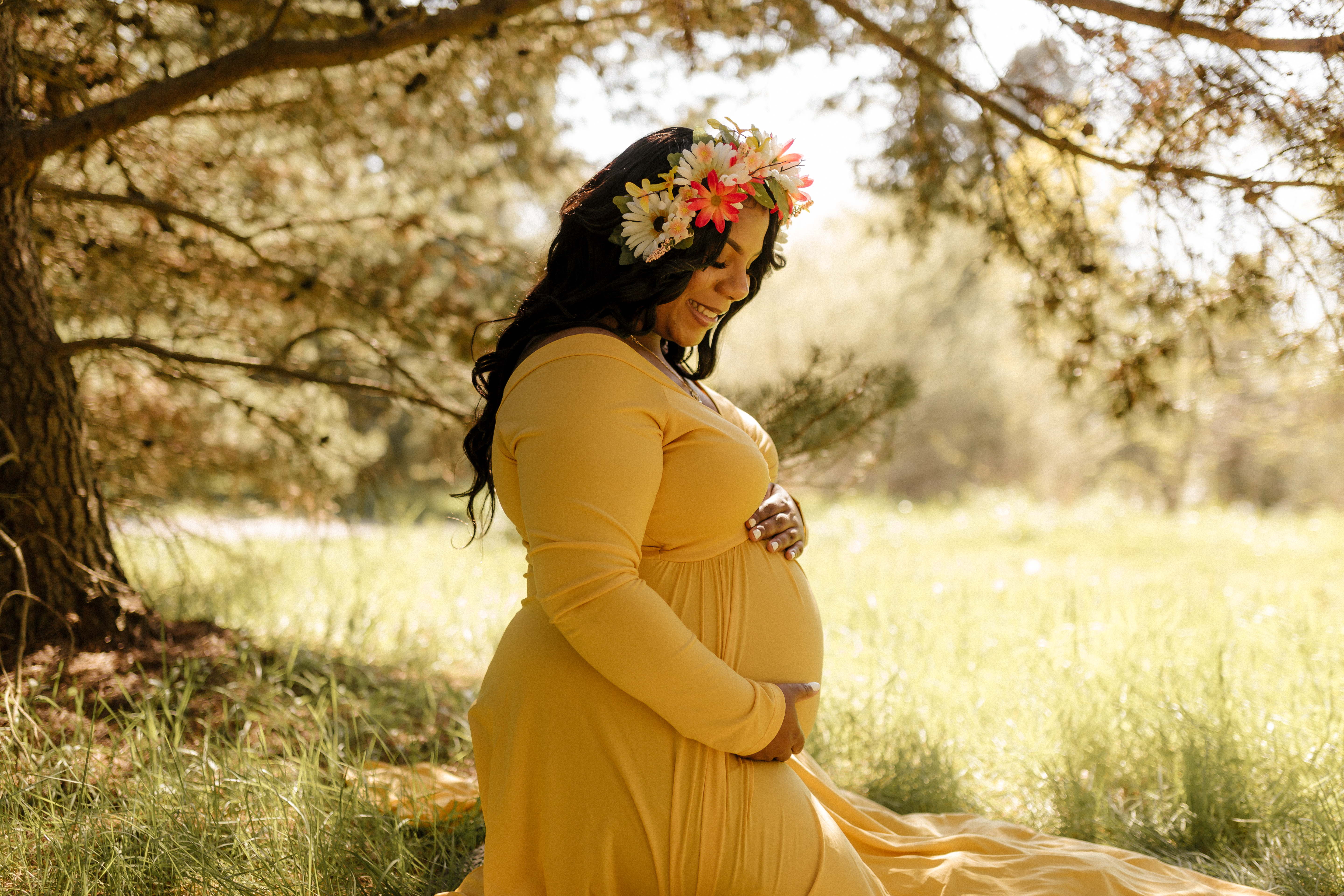 Smiling pregnant woman in a yellow dress and flower crown cradling her baby bump under sun-dappled trees, captured during a Shoott maternity photoshoot.