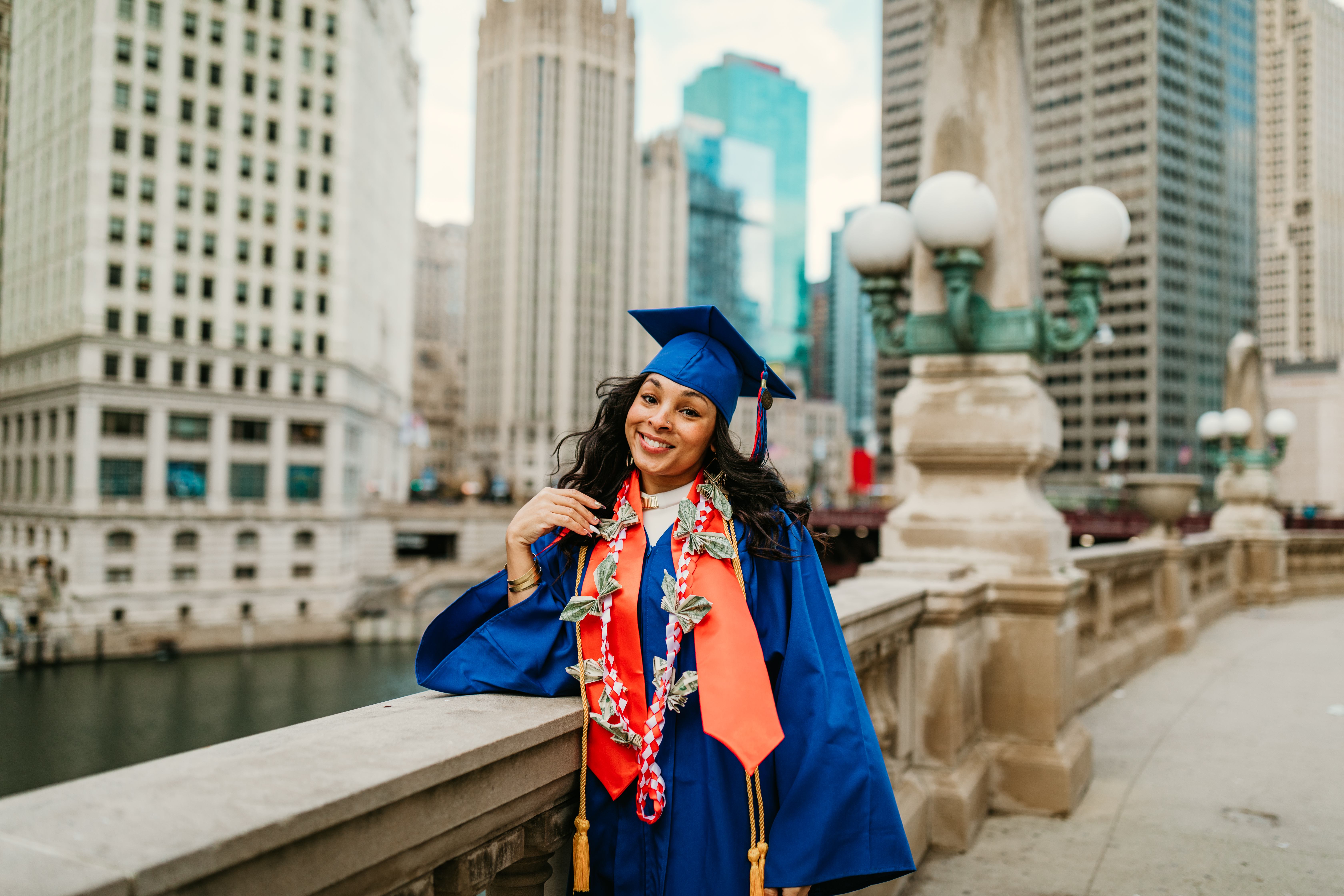 Graduate in blue cap and gown wearing a money lei on a bridge with city skyline during free Shoott photo session in Atlanta