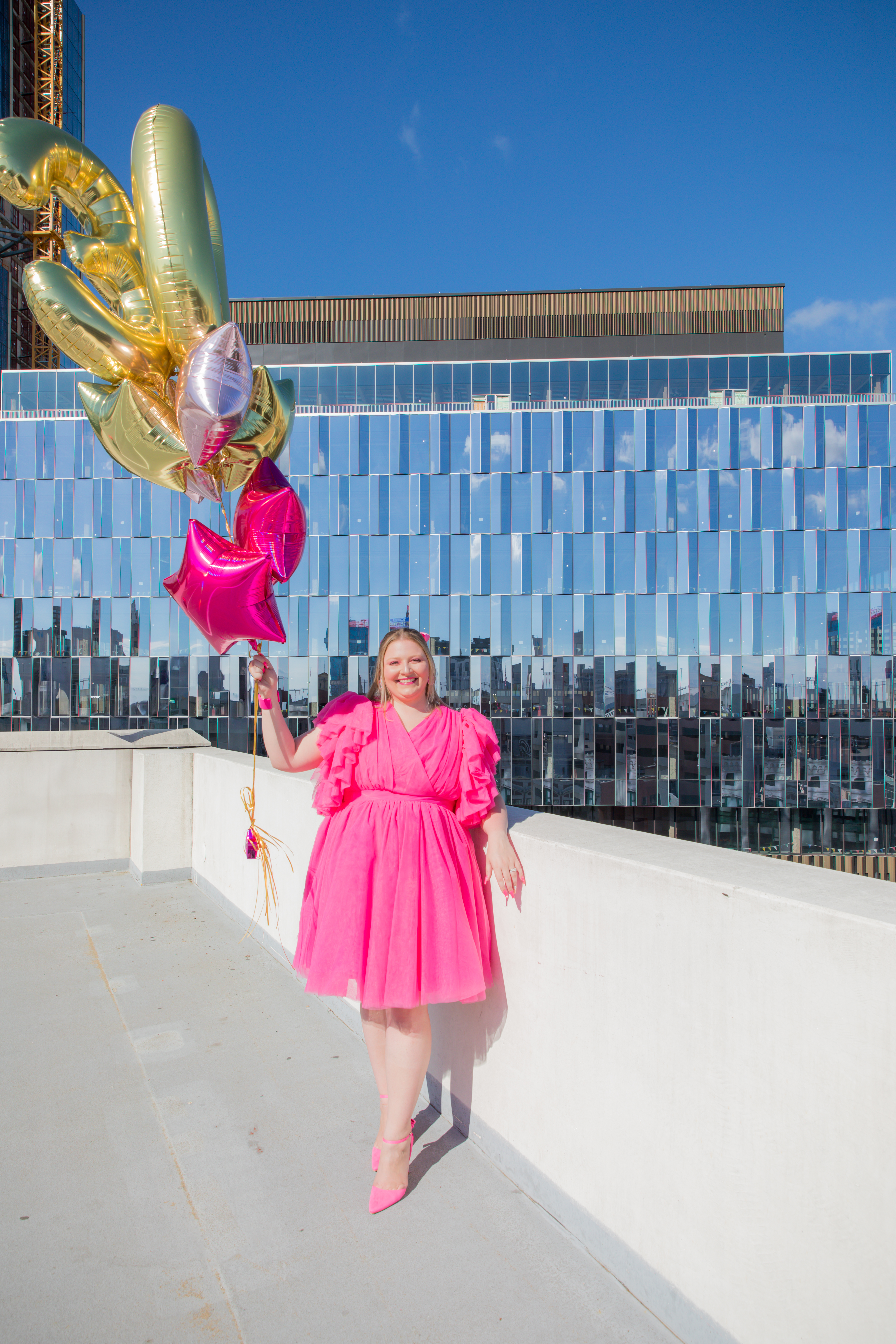 Woman in a hot pink ruffled dress smiling on a rooftop with city skyline views, holding pink and gold star balloons during her Shoott session.