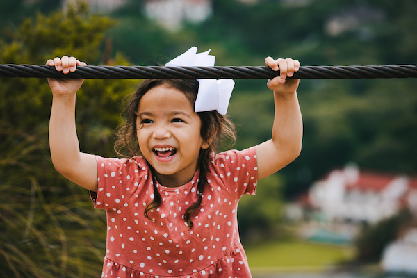 A young girl hanging from a rope and smiling in an outdoor setting.