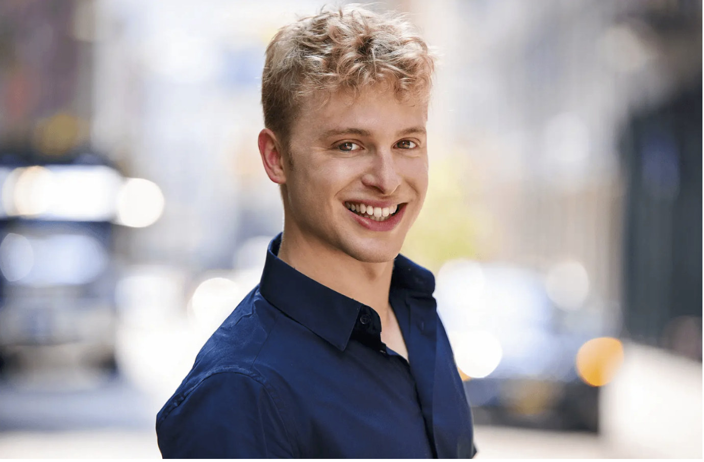 Young man with short curly blonde hair smiling outdoors in a navy blue button-down shirt, blurred city street in the background, San Antonio professional headshot by Shoott in natural light

