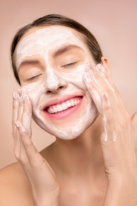 A person with a facial mask smiling with their eyes closed and hands on their cheeks.