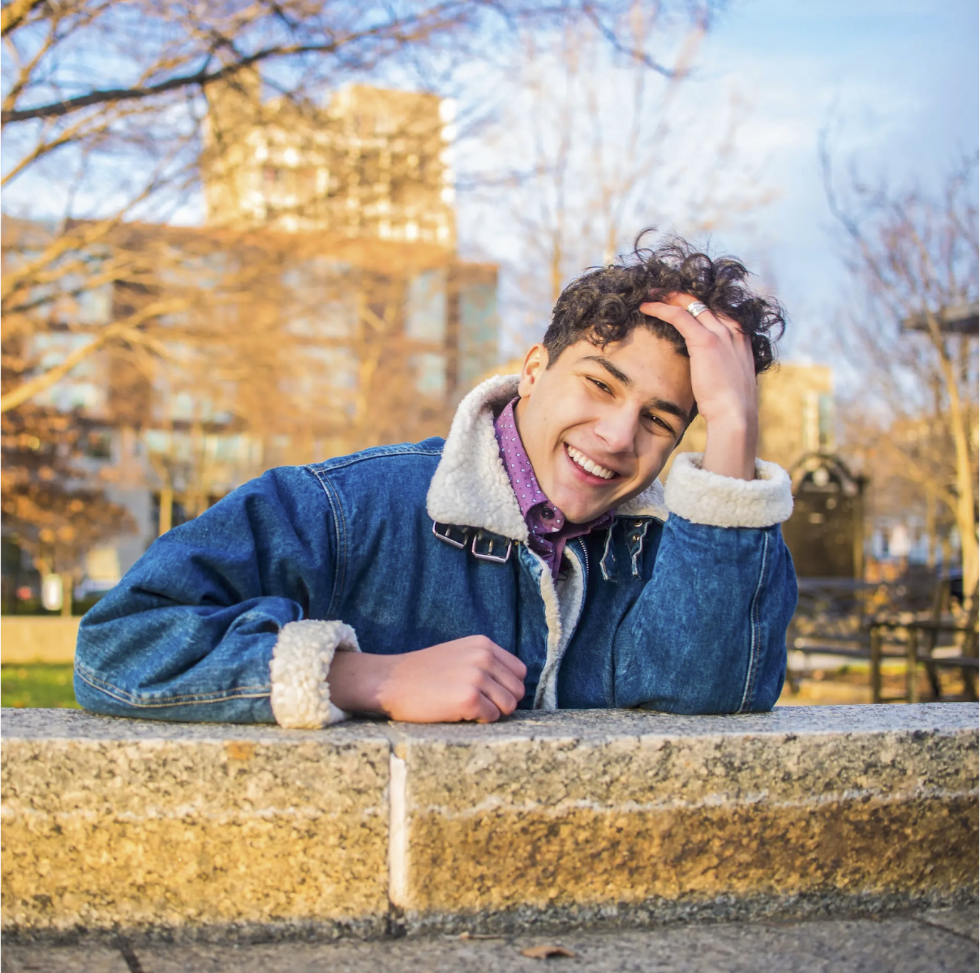 Man with curly hair laughing in a denim jacket leaning on a stone wall in a Houston park during free Shoott dating profile photo session  