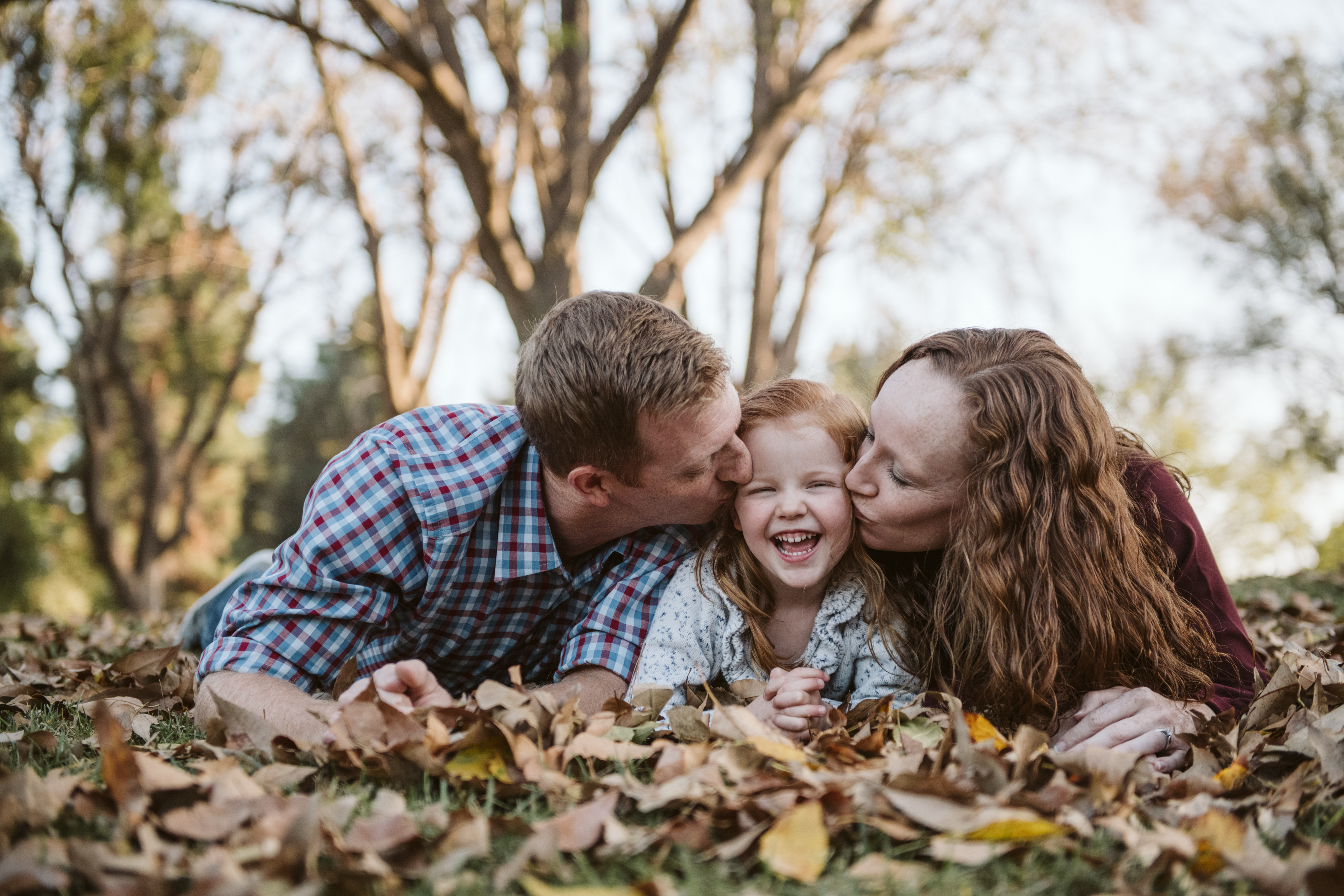 Family lying in autumn leaves as parents kiss their laughing daughter during a free Shoott lifestyle photography session
