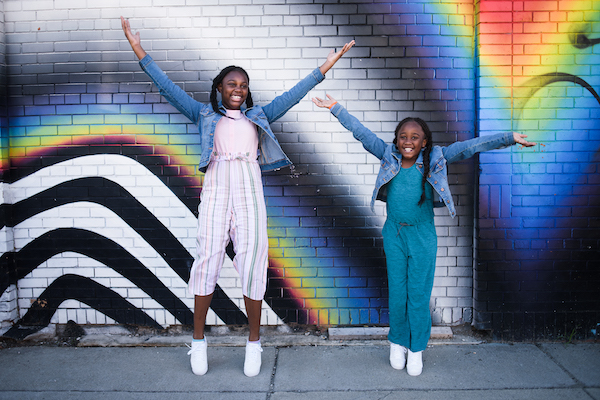 Two girls in denim jackets and jumpsuits jumping and smiling against a colorful mural