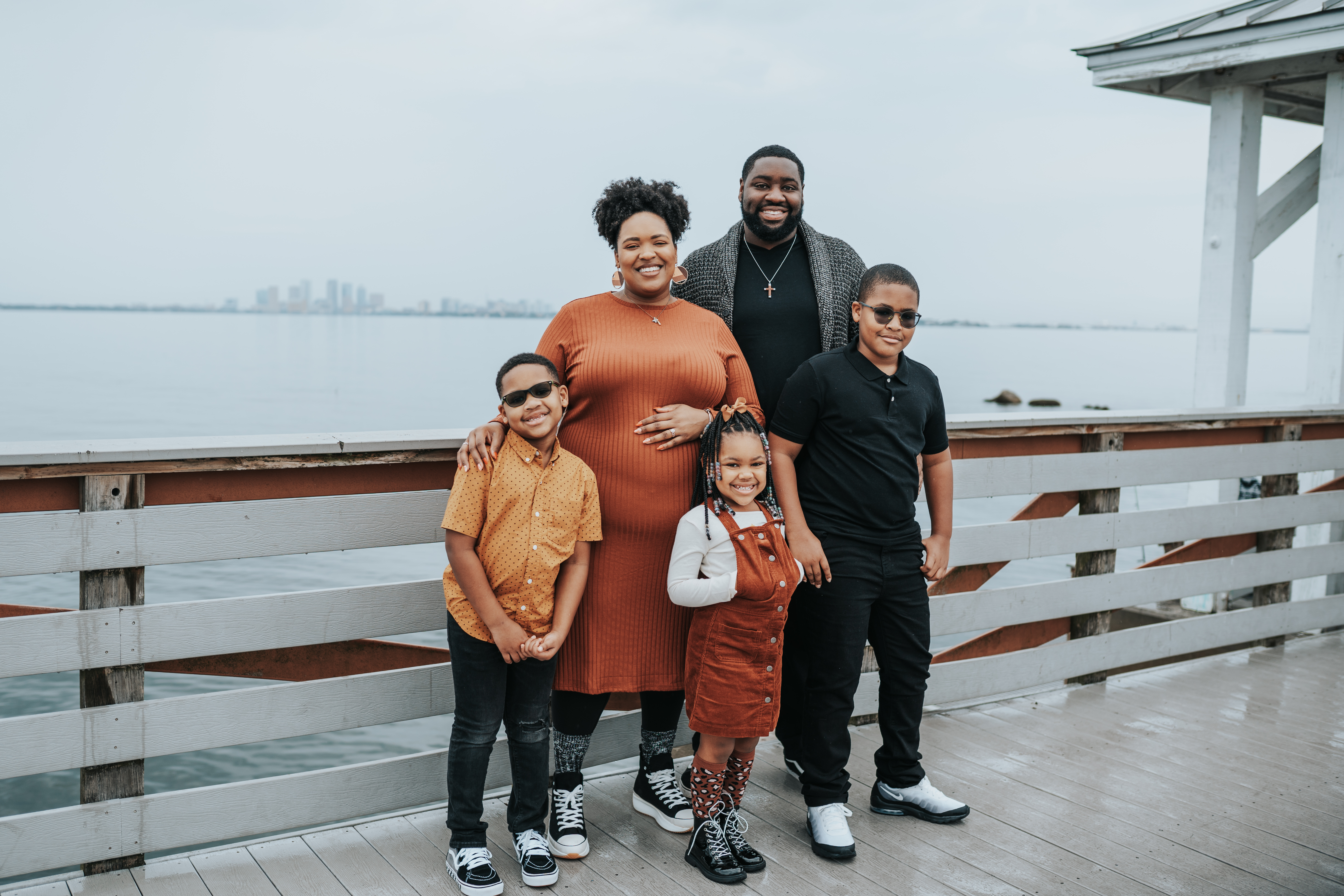 Family of five smiling on a waterfront pier with a city skyline in the background, captured during a Shoott family photoshoot.