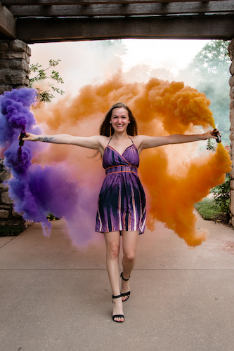 Woman standing with arms extended amidst colorful smoke grenades