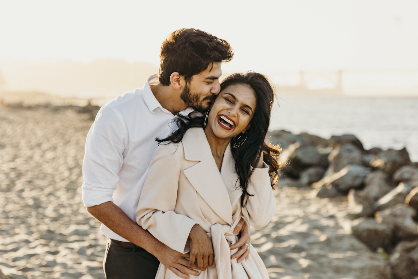 Couple laughing on a beach at sunset with him kissing her cheek during free professional lifestyle photo session with Shoott in Houston   
