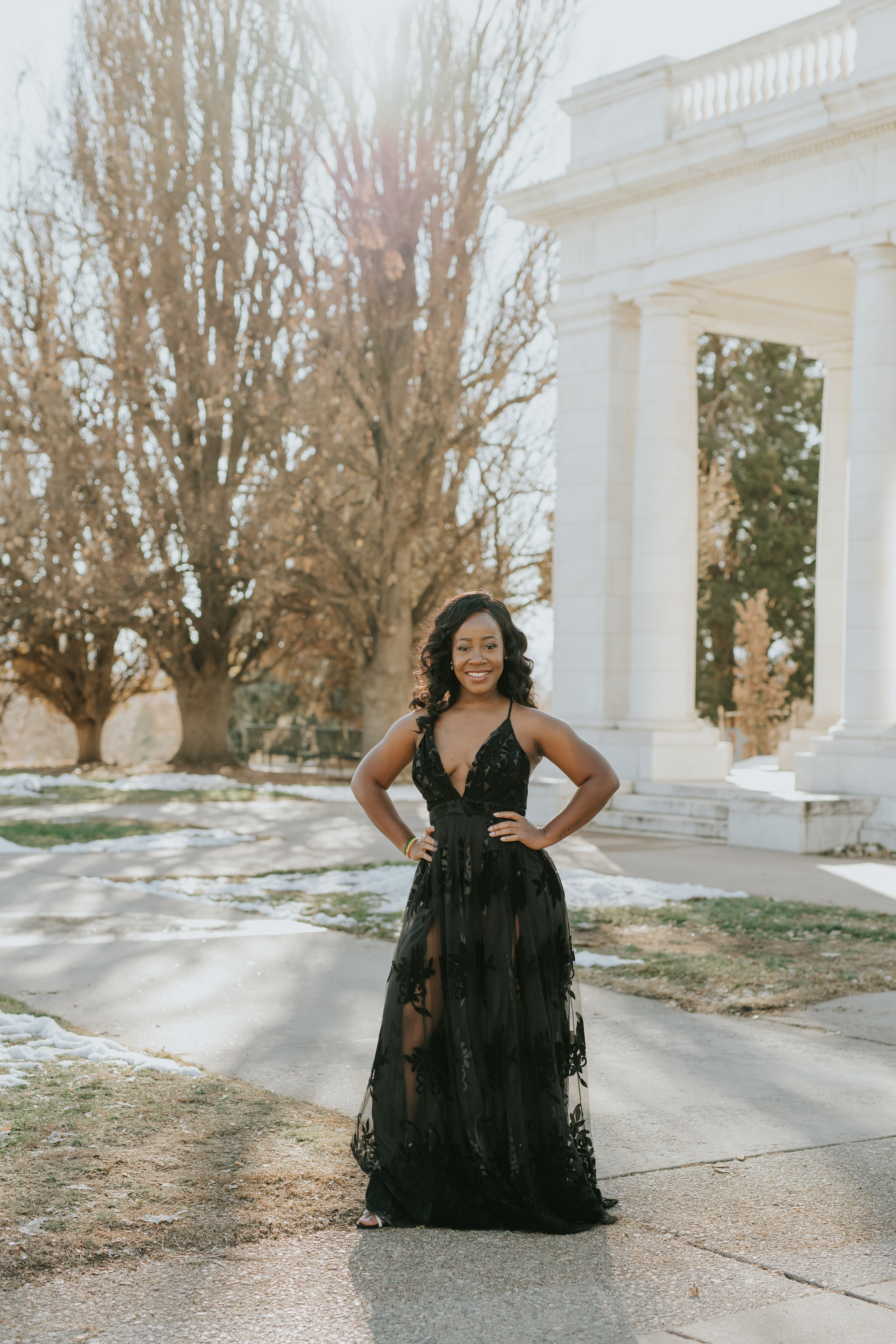 Woman in a black floral tulle gown smiling and posing with her hands on her hips in front of a white stone pavilion, one look from her Shoott session.
