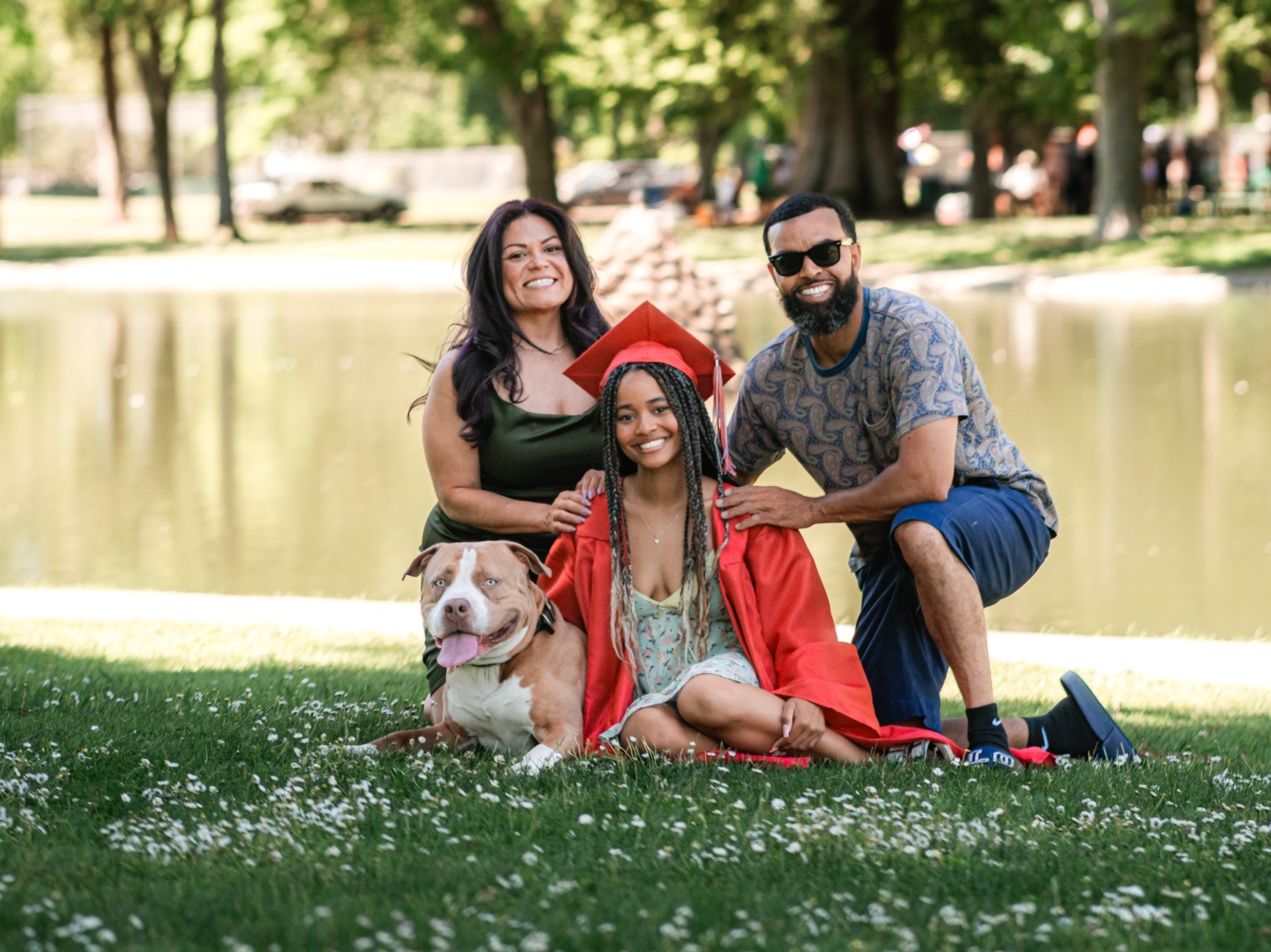 Graduate in a red cap and gown sitting on the grass by a pond with her parents and dog gathered around her, celebrating together during her Shoott session.