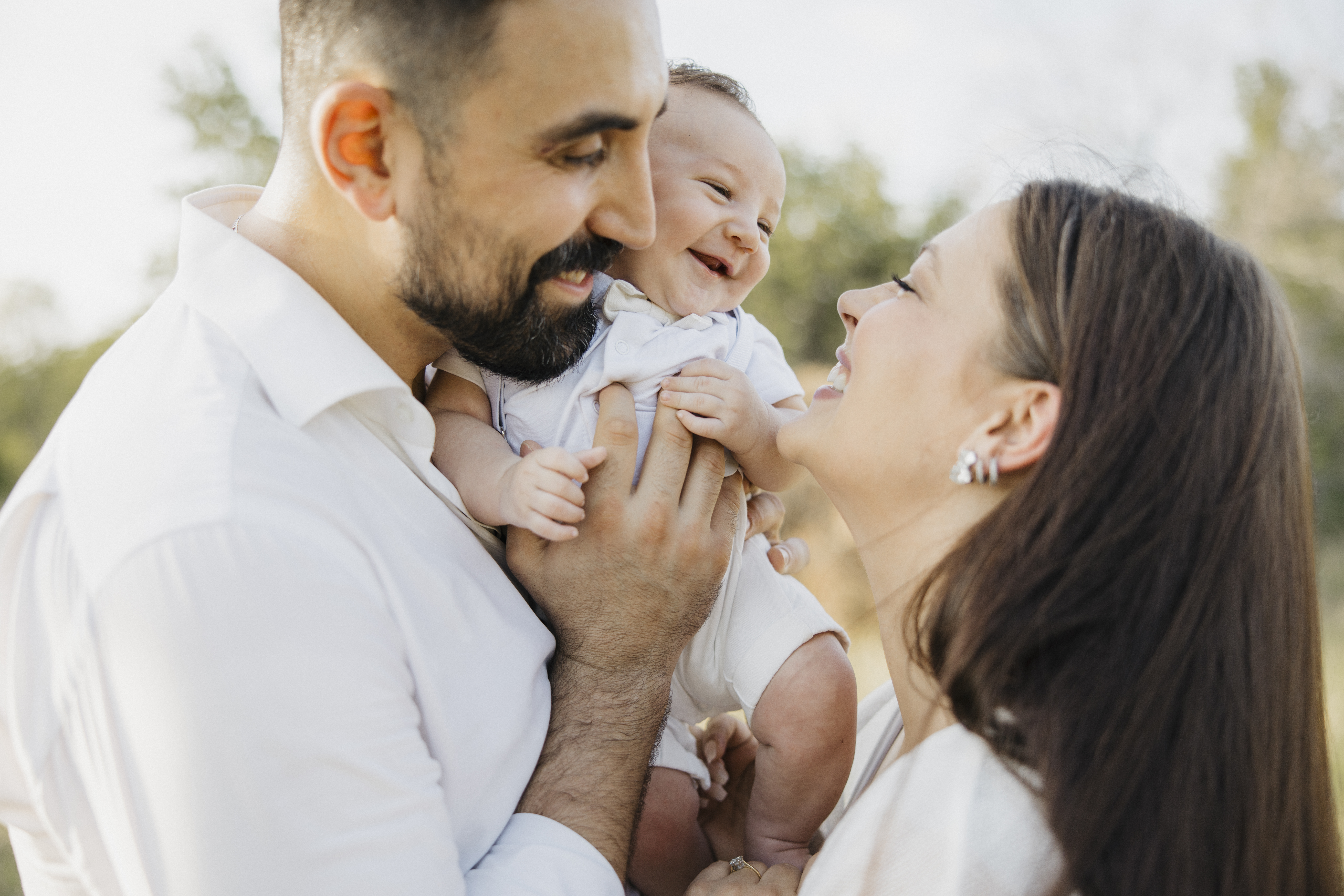 Parents in white smiling at their giggling baby held between them outdoors, captured during a Shoott family photoshoot.