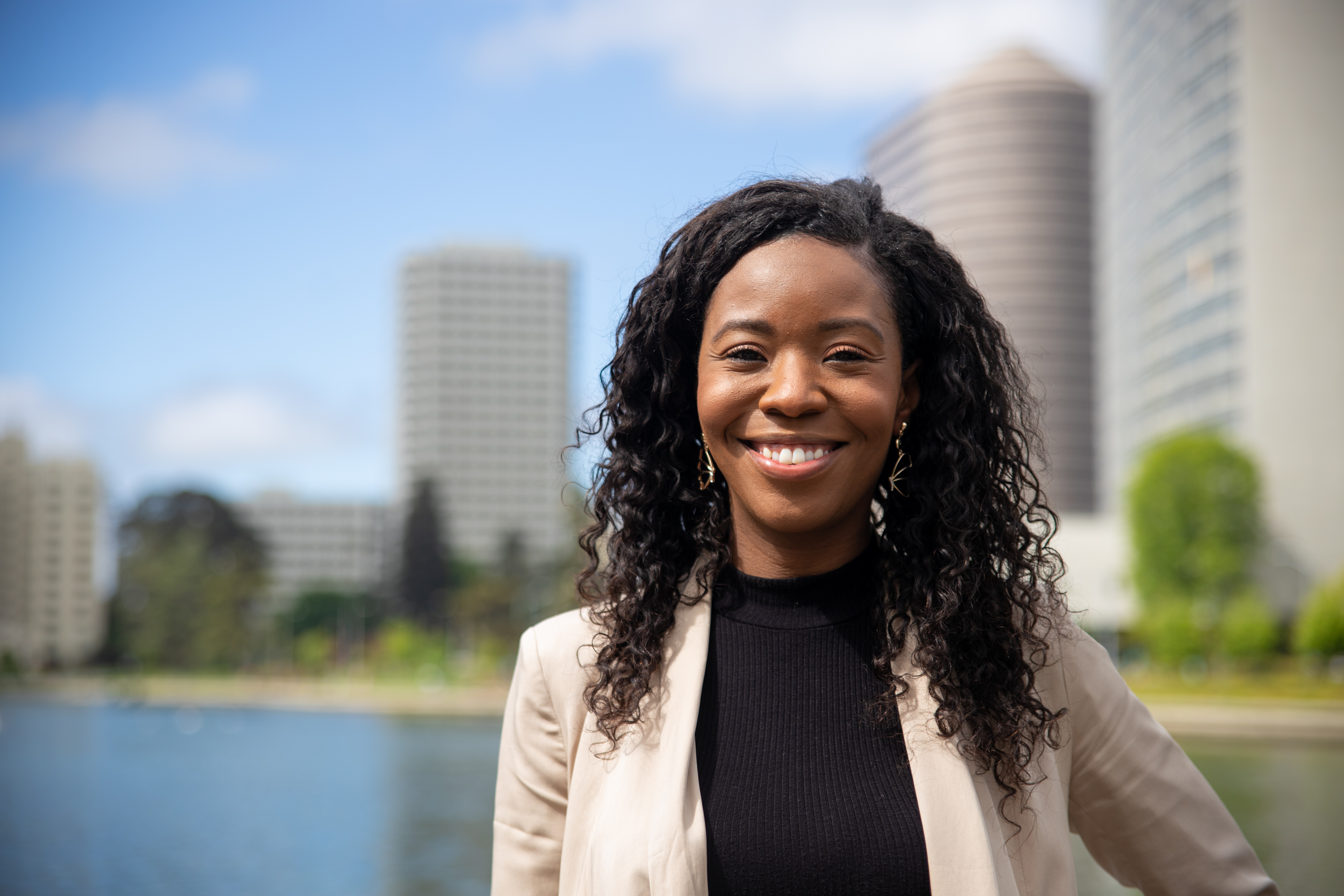 Beautiful Black woman with long curly hair smiling outdoors in a cream blazer and black top, waterfront city skyline in the background, San Antonio professional headshot by Shoott in natural light
