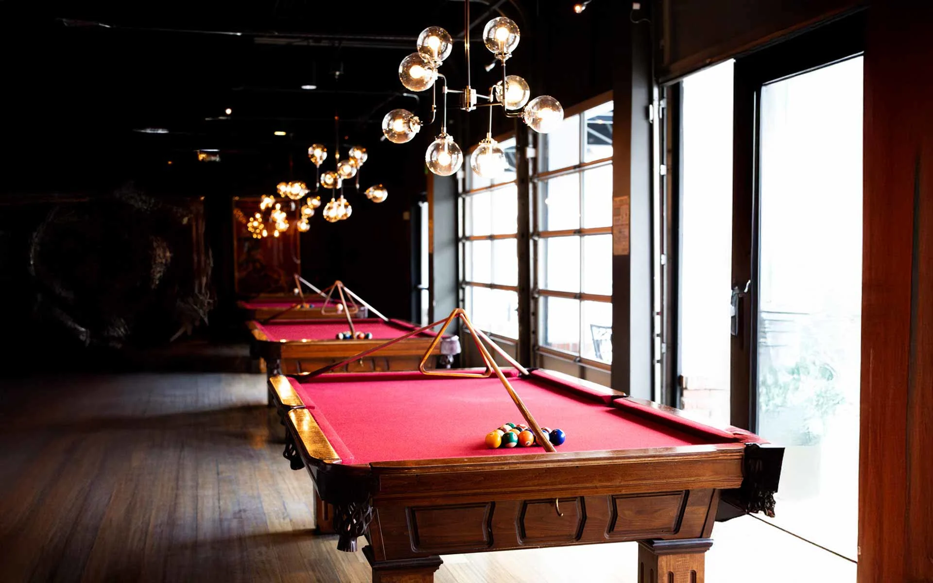 Row of red felt pool tables with overhead globe lighting inside a stylish Lucky Strike billiards lounge with natural light from large windows.