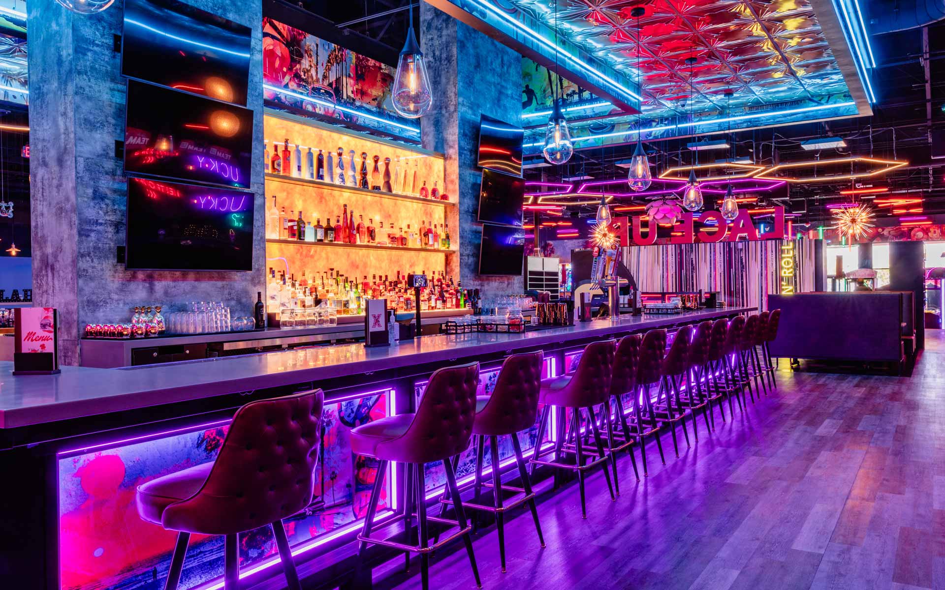 Upscale bowling lounge interior with neon lighting, illuminated bar stocked with bottles, velvet bar stools, and modern arcade-style ambiance at a Lucky Strike venue