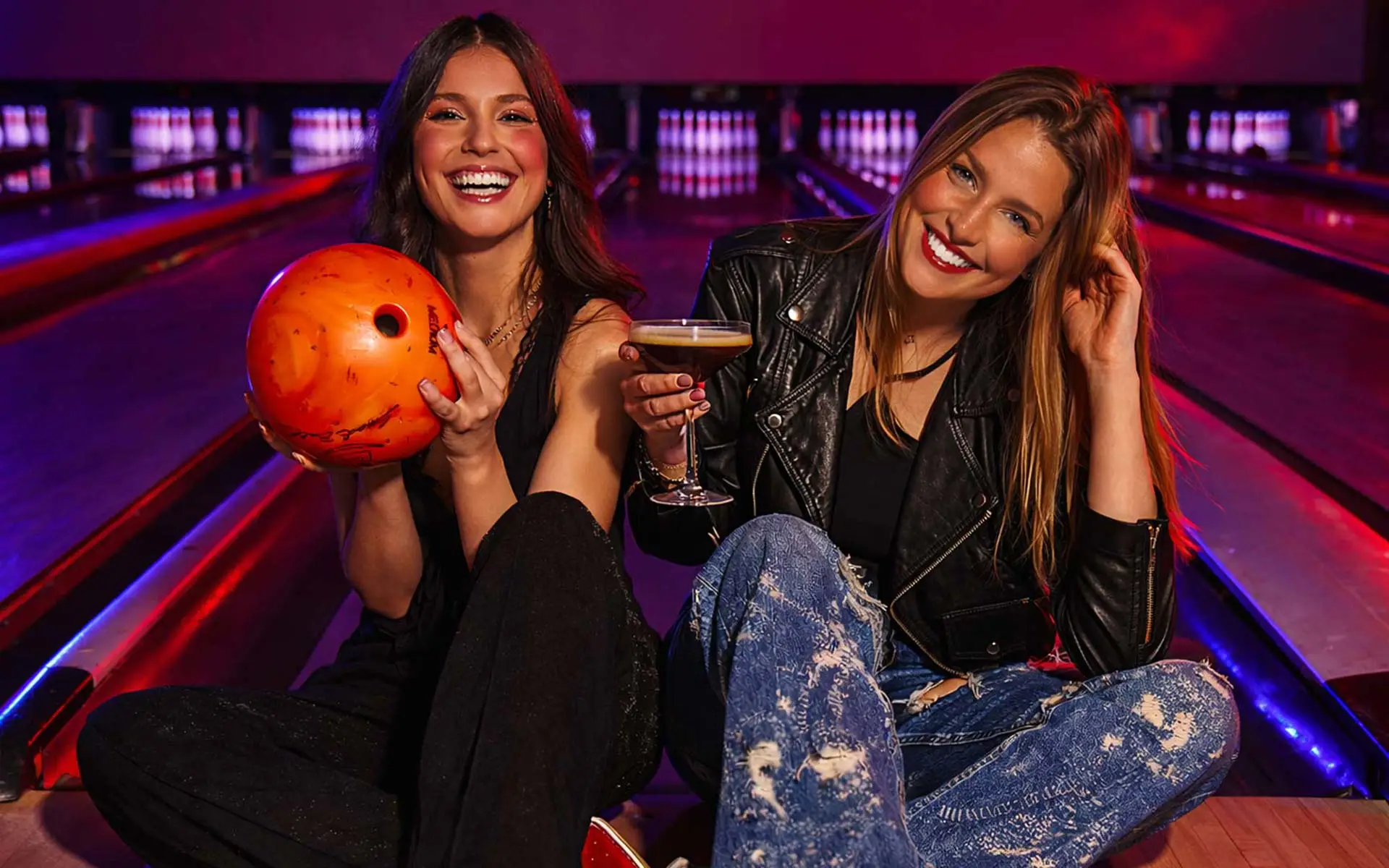 Two women sitting on a bowling lane at Lucky Strike, one holding an orange bowling ball and the other holding a cocktail, smiling under colorful neon lane lighting.