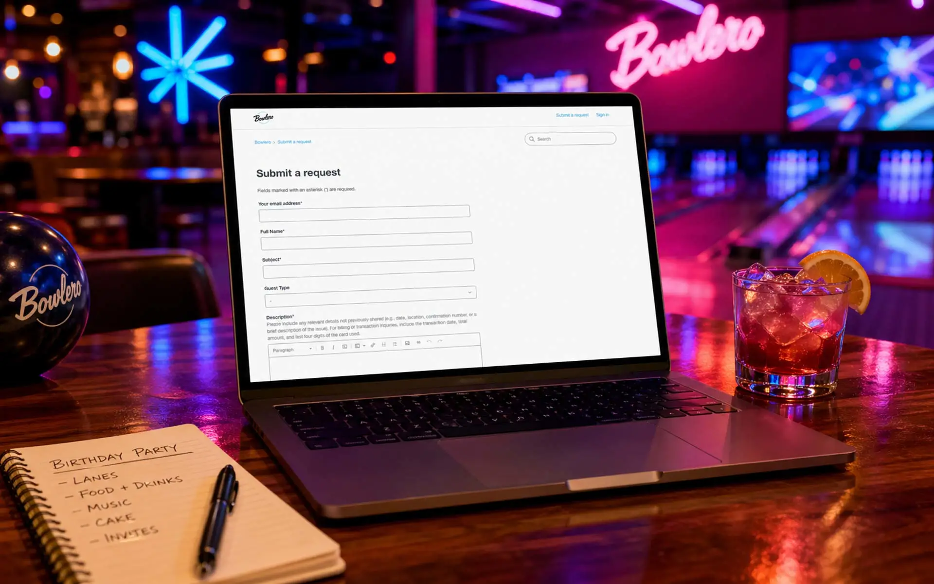 Laptop displaying a “Submit a Request” form on a glossy table inside a neon-lit Bowlero bowling alley, with a cocktail, notebook, and bowling ball nearby.