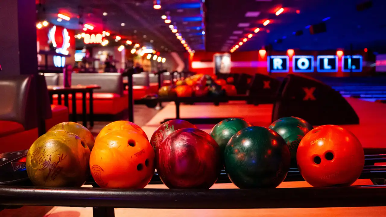 Colorful bowling balls in a return rack with bowling lanes and a large bowling center in the background.