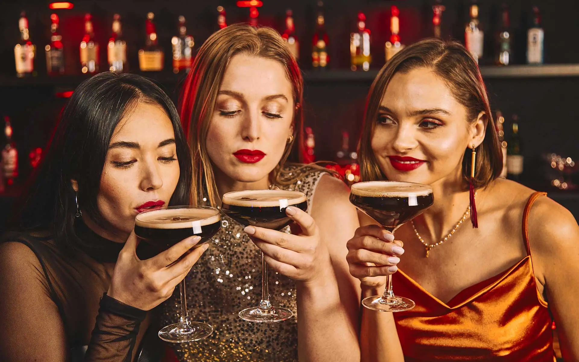 Three women enjoying espresso martinis at a stylish bar, dressed up for a night out with warm lighting and shelves of liquor bottles in the background.