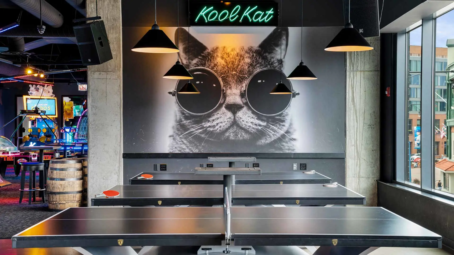 Modern game lounge with ping pong tables beneath pendant lights, featuring a large black-and-white mural of a cat wearing sunglasses and a neon “KoolKat” sign above; arcade games glow in the background and city views are visible through tall windows.