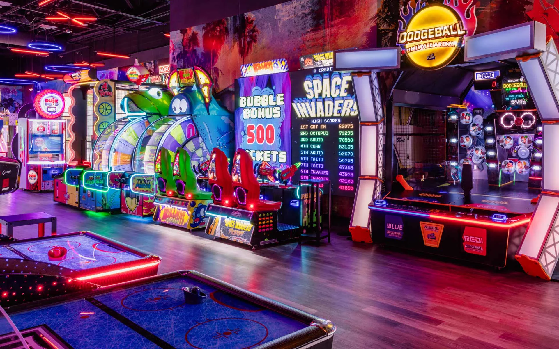 Colorful arcade area with neon lighting, featuring classic and modern game machines including air hockey, racing games, and Space Invaders in a lively Lucky Strike entertainment venue.