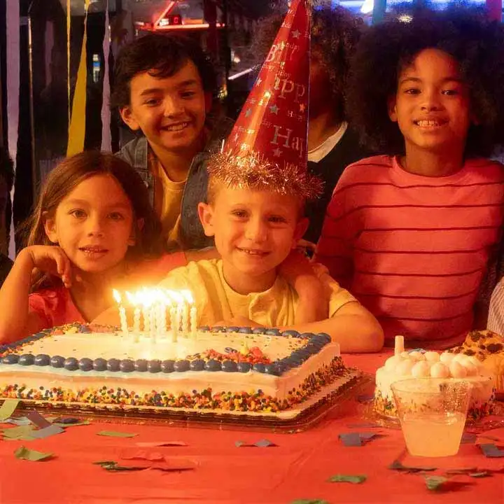 Group of children gathered around a birthday cake with lit candles at an arcade party, smiling and celebrating among colorful decorations, cupcakes, and glowing game machines in the background.