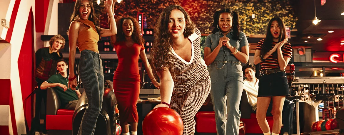 Woman rolling a bowling ball down the lane while a group cheers behind her.