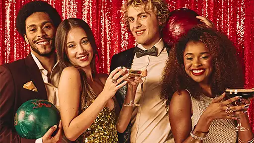 Group of friends enjoying a holiday night out at Lucky Strike, holding cocktails and bowling balls against a sparkling red backdrop.