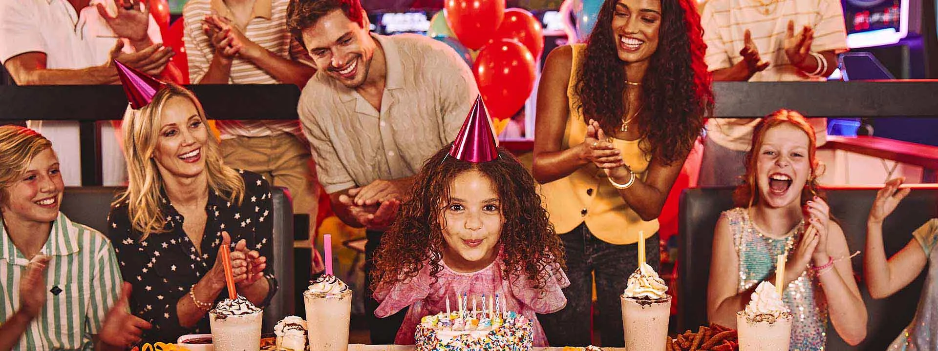 A girl with a birthday cake with candles and a large group celebrating her birthday.